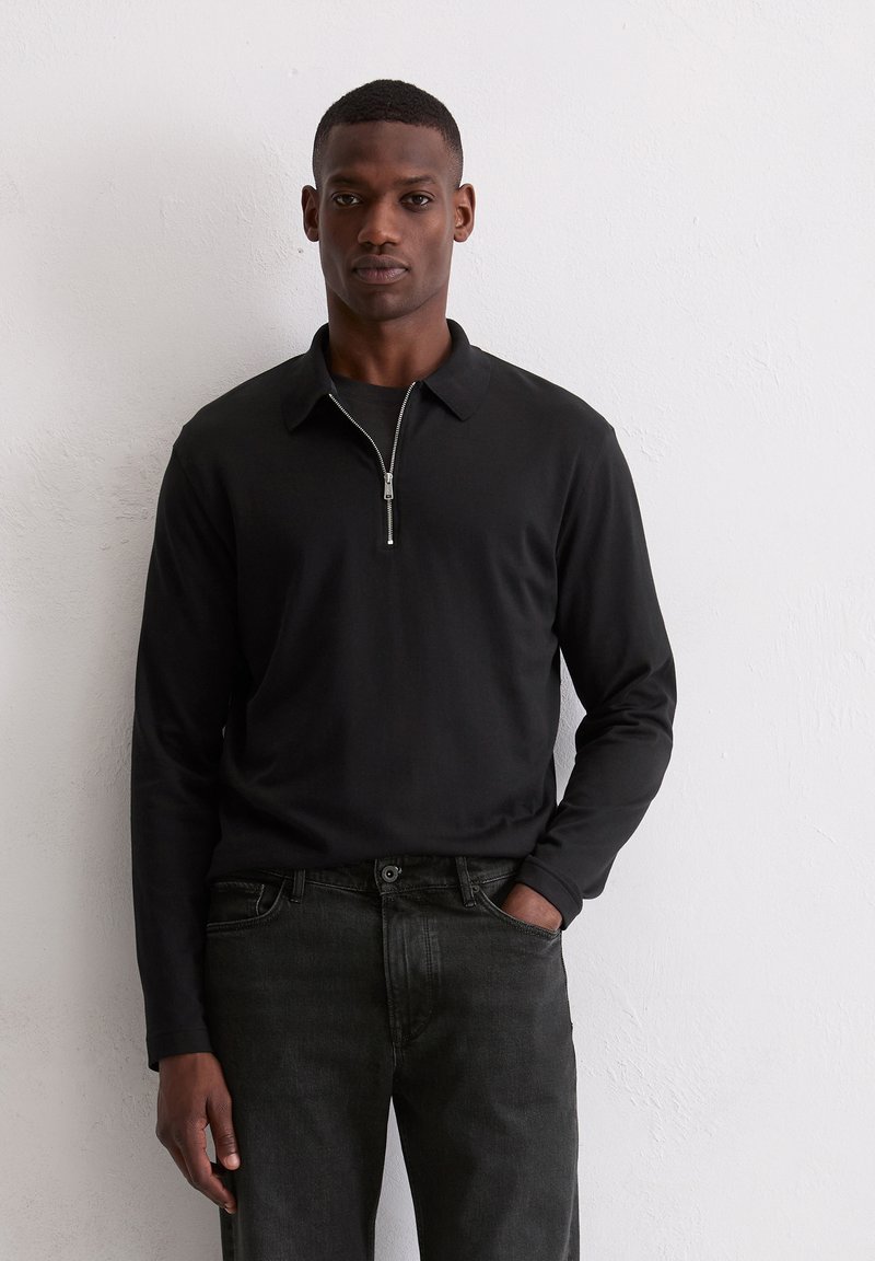 Zwart poloshirt met lange mouwen en een ritssluiting bij de kraag, gecombineerd met donkere jeans. Gladde textuur en nauwsluitend ontwerp, geschikt voor casual draagstijl.