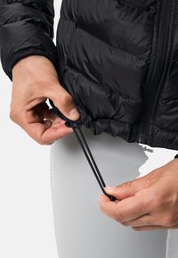 Zwarte pufferjack met een elastisch trekkoord aan de onderkant. Hand die het trekkoord vasthoudt met een soepele textuur. Grijze leggings zichtbaar eronder.