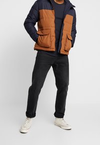 Męska kurtka puchowa z granatową górą i miedzianym dołem, z dwoma przednimi kieszeniami i zamkiem błyskawicznym. Noszona z czarnymi dżinsami i białymi trampkami.