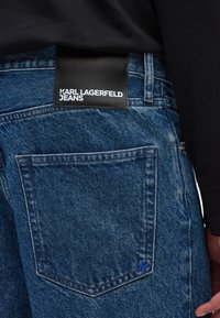 Tumed sinised teksad, millel on must nahast silt, millel on kirjas "KARL LAGERFELD JEANS" ja õmmeldud sinine logo taskul.