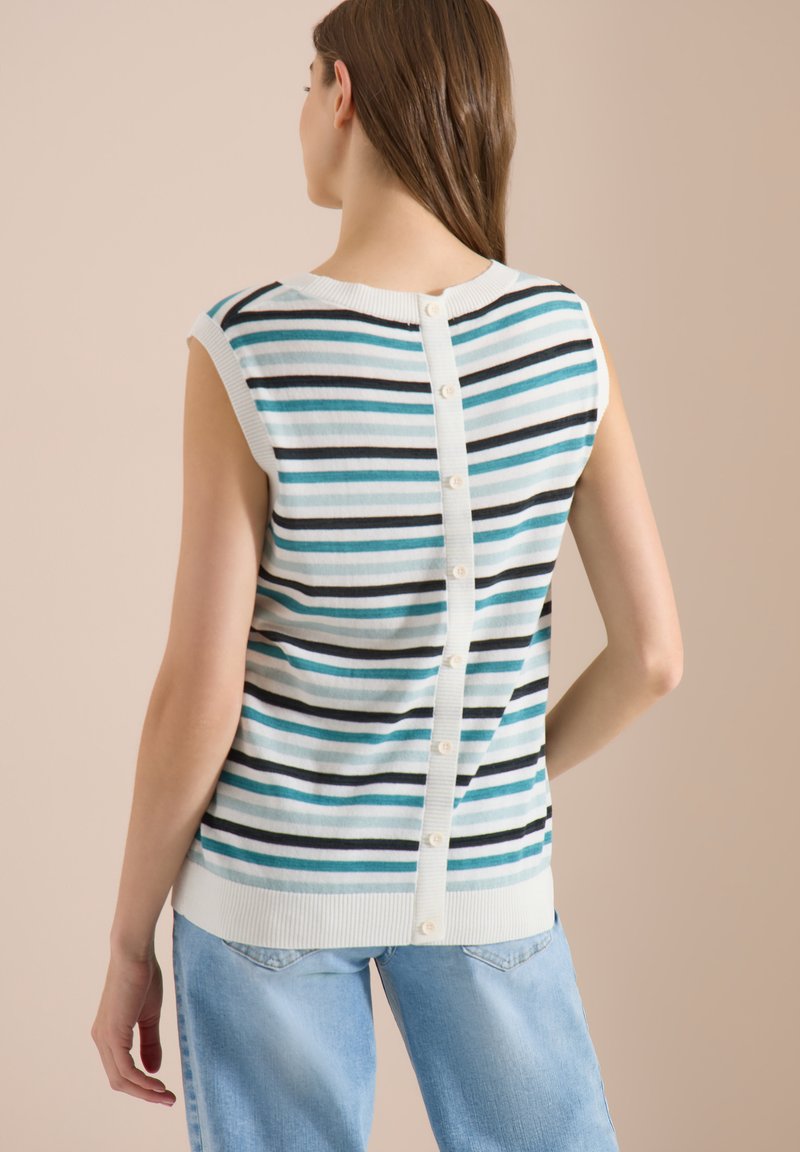 Femme portant un pull rayé sans manches avec un détail de boutons dans le dos et un jean bleu clair, debout de dos à la caméra.