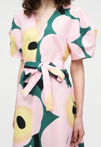 Γυναίκα που φοράει ροζ φόρεμα με μεγάλα κίτρινα και πράσινα λουλούδια, κοντά μανίκια, V-λαιμόκοψη και ζώνη μέσης δεμένη με φιόγκο.