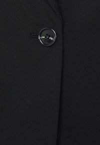 Czarna tkanina o fakturowanym wykończeniu, z widocznym czarnym plastikowym guzikiem po boku oraz detalem wykończenia klapy z precyzyjnym szwem.