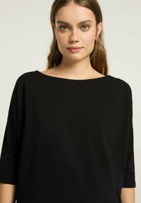 Czarna luźna bluzka z szerokim dekoltem w łódkę i rękawami o długości trzy czwarte, wykonana z gładkiego materiału, charakteryzująca się minimalistycznym designem.