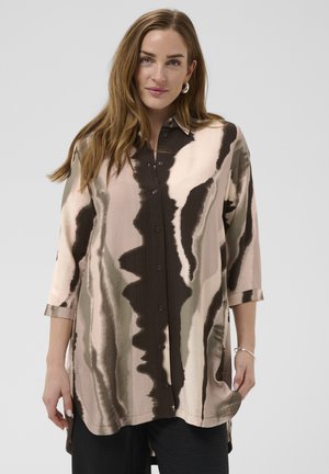 Woman with long brown hair wearing a loose, buttoned shirt with abstract brown, beige, and olive patterns and black pants against a plain background.