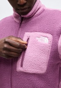 Person iført en lilla fleecejakke med The North Face-logo, som lynlås et lille brystlomme med deres højre hånd.