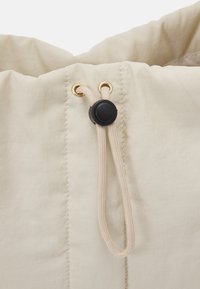 Tissu beige avec coutures cousues, featuring un cordon beige passé à travers des œillets dorés avec un bloque-cordon en plastique noir.