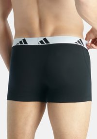 Boxer shorts pretos com uma cintura elástica branca, apresentando detalhes do logótipo preto da Adidas. Tecido suave com um design ajustado.