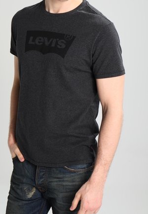 Mężczyzna w ciemnoszarej koszulce Levi's i niebieskich jeansach, stojący z jedną ręką w kieszeni na jednolitym jasnym tle.