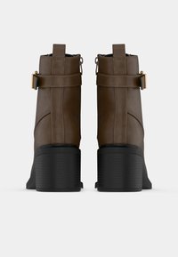 Καφέ μποτάκια μέχρι τον αστράγαλο από δέρμα, με υφή και μαύρη ελαστική φούστα. Διαθέτουν φερμουάρ στο πλάι, λεπτομέρειες με λουριά και κατακόρυφες ραφές.