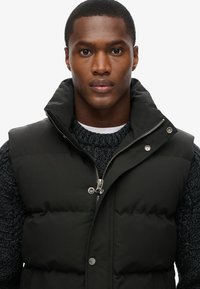Chaleco acolchado negro con cuello alto, cierre delantero con cremallera y botones a presión. Lleva puesto sobre un suéter oscuro texturizado, mostrando un look en capas.