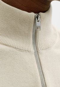 Gros plan d'un pull beige en maille côtelée avec une fermeture éclair argentée partiellement fermée près du cou, sur un fond de peau foncée.