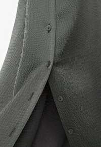 Chemise en tissu texturé vert foncé avec de petits boutons noirs ; comporte un motif en relief subtil et une fente latérale pour faciliter le mouvement.