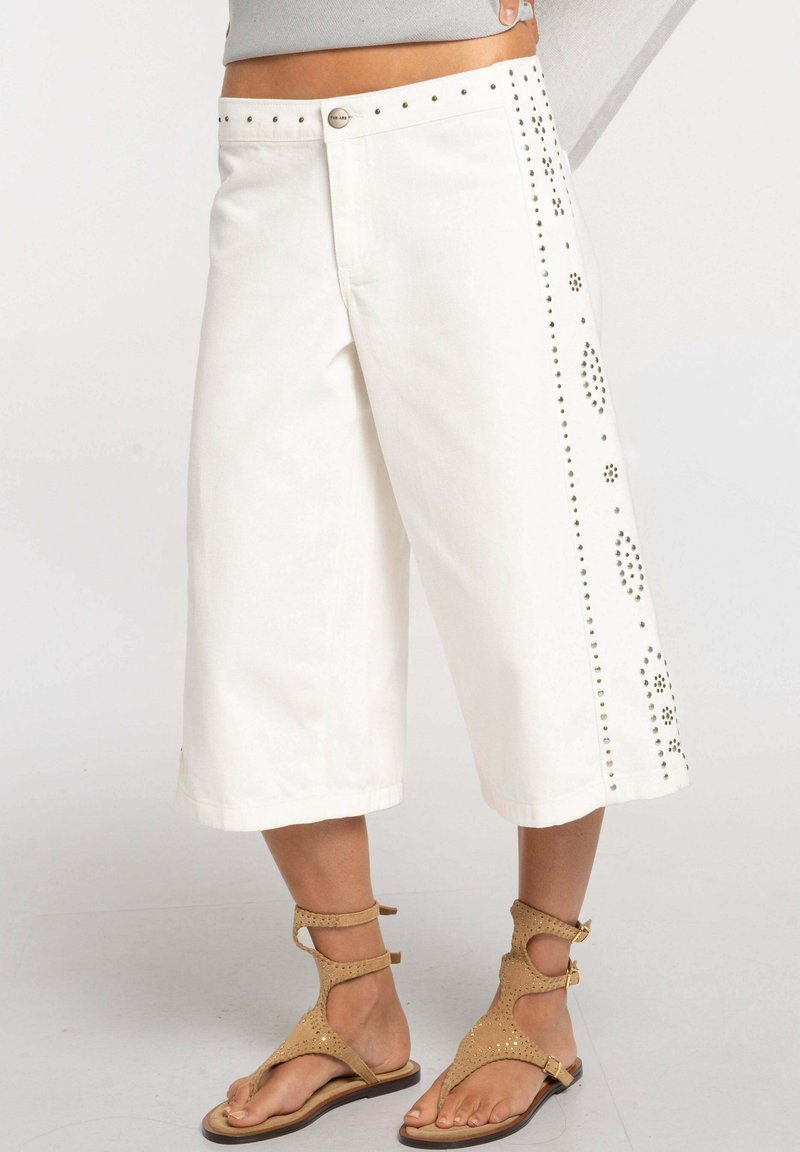 Pantalones blancos de pierna ancha y corta con patrones florales tachonados en los laterales, usados con sandalias planas marrones con tiras.