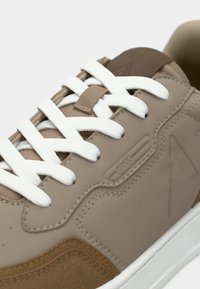 Béžové tenisky s bílými tkaničkami, hladkým syntetickým svrškem, texturovanými akcenty, semišovými detaily a gumovou podrážkou. Minimalistický design.