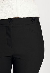 Pantalons noirs avec une texture de tissu lisse, deux poches avant et deux boutons noirs à la taille. Détails de surpiqûre accentués.