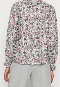 Blusa floreale di colore azzurro chiaro, con motivi multicolore in rosso, marrone e blu navy. Maniche lunghe con polsini arricciati e vestibilità leggermente ampia.