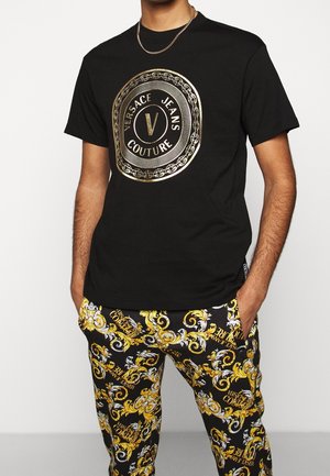 Zwart katoenen T-shirt met een groot gouden cirkelvormig logo met "Versace Jeans Couture." Draagt bijpassende zwarte en gele broek met patroon.