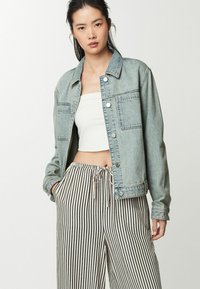 Giacca di jeans blu chiaro con due tasche sul petto, chiusura con bottoni e vestibilità leggermente oversize, abbinata a pantaloni larghi a righe bianche e nere.