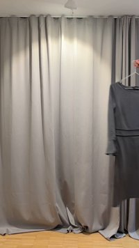 Ruka drží čierne šaty s dlhým rukávom na vešiaku pred svetlosivými závesmi od podlahy po strop a drevenou podlahou.