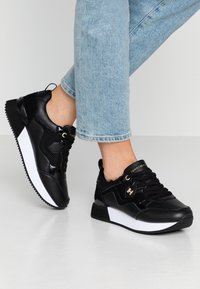 Baskets de sport noires avec un mélange de cuir lisse et de mesh, dotées d'une semelle intermédiaire blanche, de lacets ronds et d'accents décoratifs en métal.