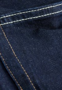 Tumši zilas džinsu audums ar redzamām dūrēm kontrastējošā gaiši brūnā krāsā. Materiāls rāda vieglu tekstūru un graudu. Tuvplāns.