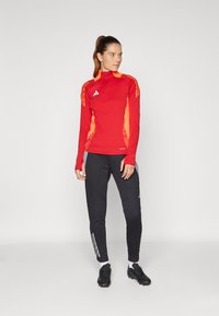 Rotes und oranges langärmliges Sportoberteil mit halbem Reißverschluss, kombiniert mit schwarzen schmal geschnittenen Hosen mit weißen Seitenstreifen und schwarzen Fußballschuhen.