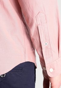 Ljust rosa långärmad skjorta med knappdetaljer, gjord av texturerat tyg. Har en spetsig ok och rundad nederkant, tillsammans med marinblå byxor.