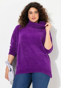 Lila oversized polokrage tröja i mjukt, texturerat material med ribbade mudd och nederkant. Bärs med en ljusgrå väska och jeans.