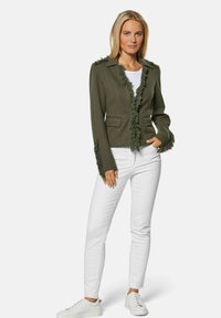 Olijfgroen blazer met rafeldetails bij de kraag en manchetten, gecombineerd met witte, strakke jeans en witte sneakers. Gladde textuur, gestructureerd ontwerp.