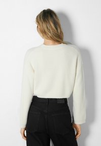 Weißer, verkürzter Pullover mit strukturiertem Finish und geripptem Ausschnitt, kombiniert mit hochgeschnittenen schwarzen Hosen. Der Stoff wirkt weich und warm.