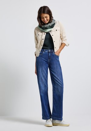 Giacca di velluto a coste beige, top verde scuro, jeans blu a gamba larga e sciarpa a righe verde e nera, indossata con sneakers bianche.