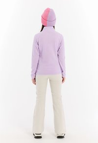 Lichtpaarse fleecjacket met een hoge kraag, gecombineerd met crèmekleurige broek. Een roze en lavendel gebreide muts maakt de outfit compleet.