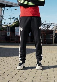 Čierne teplákové nohavice Adidas s bielymi pruhmi, obsahujúce nemecké futbalové logo a tri hviezdy. V kombinácii s čierno-bielou športovou obuvou.