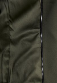 Olivově zelená textilie s hladkou strukturou, na které se nachází kontrastní černý zip s malými stříbrnými detaily podél švu.