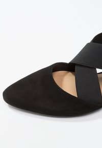 Chaussure plate noire à bout pointu avec des brides élastiques croisées et une doublure intérieure beige sur fond blanc.