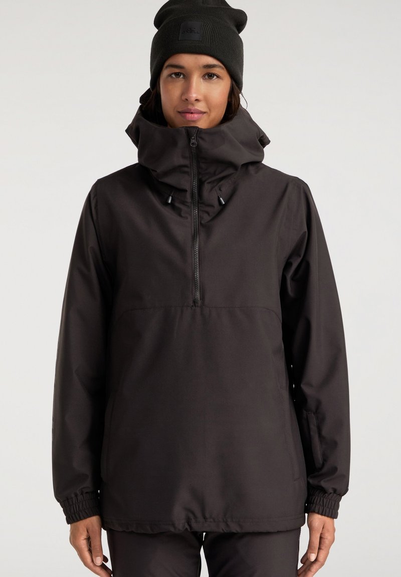 Mujer vestida con una chaqueta negra de exterior con un cuello alto y media cremallera, combinada con un gorro negro, de pie frente a un fondo liso.