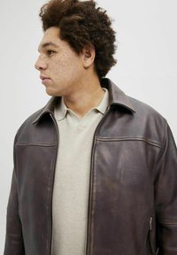 Brown leather jacket with a zip closure, stand collar, and subtle sheen. Worn over a light-colored polo shirt with a textured pattern.
