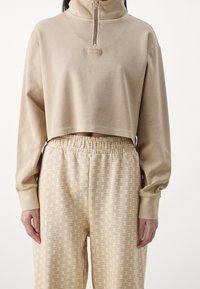 Korte beige sweatshirt met hoge kraag en rits, gecombineerd met bijpassende taps toelopende broek met een herhalend logo-patroon in lichtbruin.