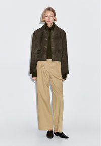 Hnědá krátká semišová bunda s bočními kapsami přes tmavě zelený rolák, doplněná širokými béžovými kalhotami a černými mules.