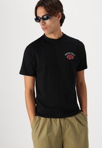 Black cotton t-shirt featuring a red and green logo design on the chest. Model wears khaki shorts and dark sunglasses.