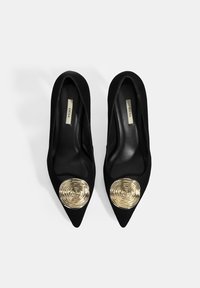 Escarpins en cuir suédé noir avec un design à bout pointu. Ils présentent un accent en spirale doré bien visible à l'avant. Doublure intérieure lisse.