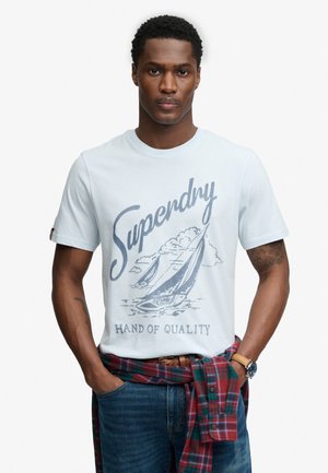 Mand iført hvid "Superdry" t-shirt med sejlbåds grafik, blå jeans, rød-grønternet skjorte bundet om taljen og ur på venstre håndled.