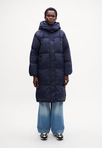 Cappotto puffer navy con cappuccio, caratterizzato da una silhouette squadrata, bottoni frontali e dettagli con cordoncino. Abbinato a jeans azzurri a gamba larga.