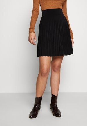 Modèle portant un pull marron ajusté et côtelé accompagné d'une jupe plissée noire, associé à des bottes de cowboy brunes brillantes jusqu'aux genoux.