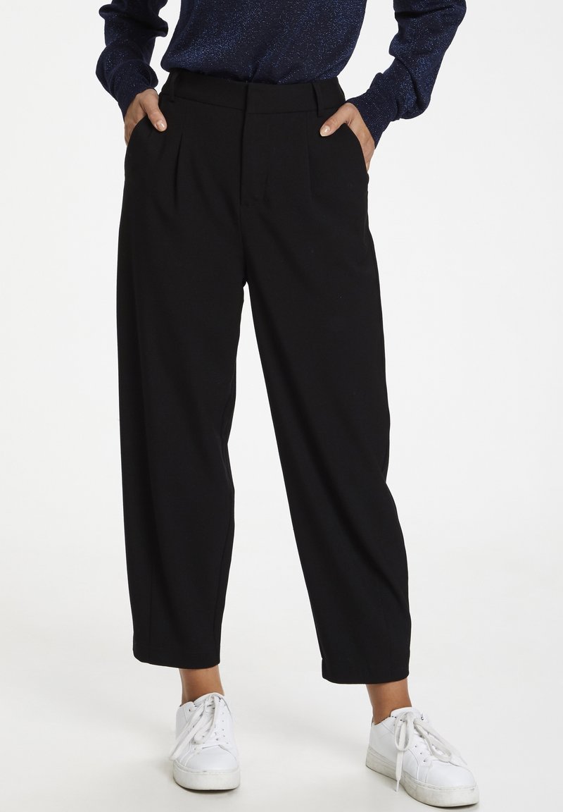Pantalon noir taille haute avec plis, coupe à jambes larges, texture lisse, poches avant, associé à des baskets blanches.