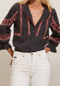 Blouse noire avec motifs brodés en rose, jaune et bleu. Présente un devant boutonné et des manches à volants. Associée à un jean blanc.