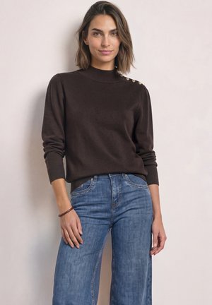 Femme portant un pull marron foncé avec des boutons dorés sur l'épaule et un jean bleu taille haute, debout contre un mur clair.