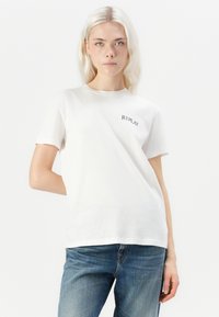 Witte katoenen t-shirt met korte mouwen, ronde hals, en 'REPLAY' in het zwart gedrukt op de linkerborst. G gedragen met blauwe denim jeans.