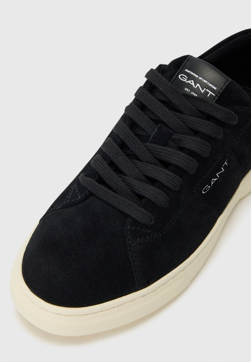Svart mocka sneakers med snörning, vit gummisula och GANT-logotyp på sidan och tungan. Slät yta med minimal design.